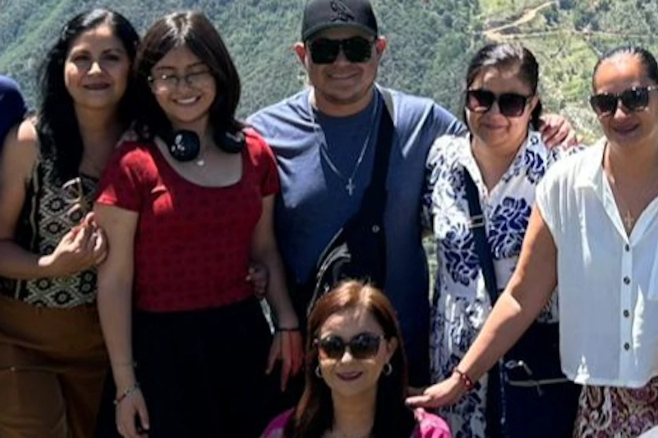 A group of eight people posing outdoors on a grassy hilltop with scenic mountains and valleys in the background under a blue sky.