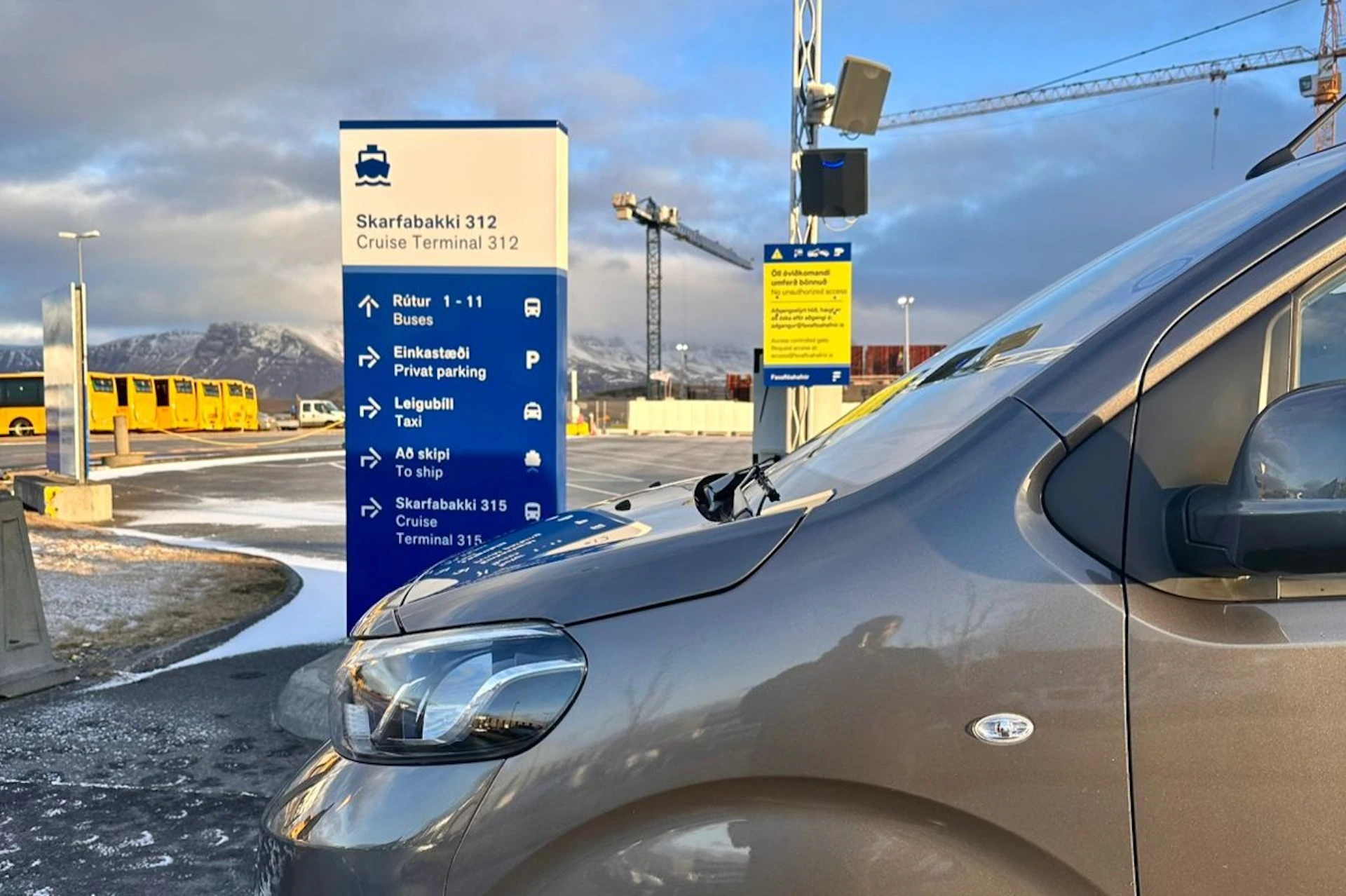 A car parked near a sign listing parking areas, bus and taxi stops, and cruise terminals. Construction cranes are visible in the background.