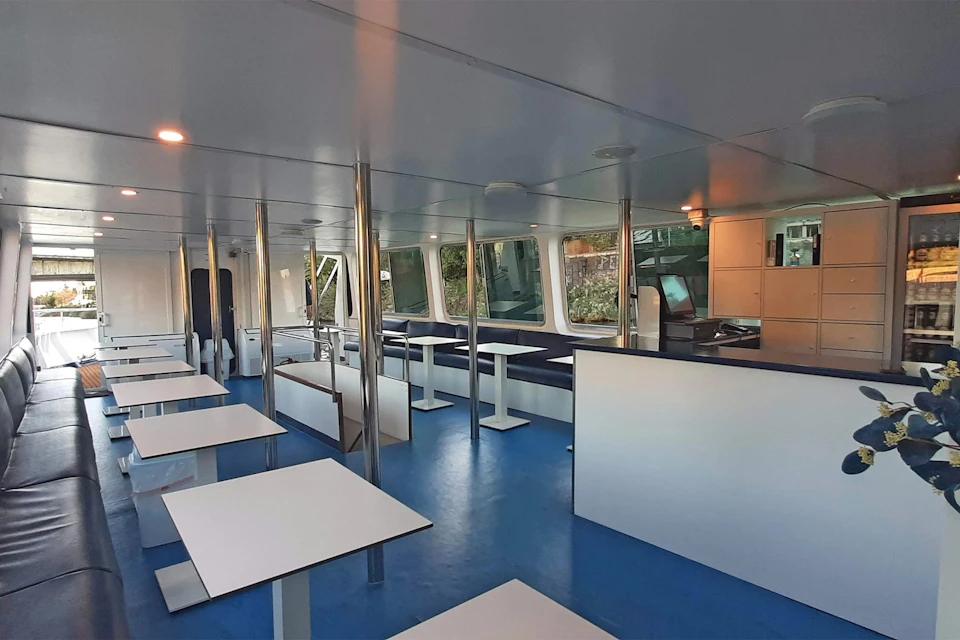 The interior of a modern ferry with blue flooring, white tables and large windows offering views of the outside.