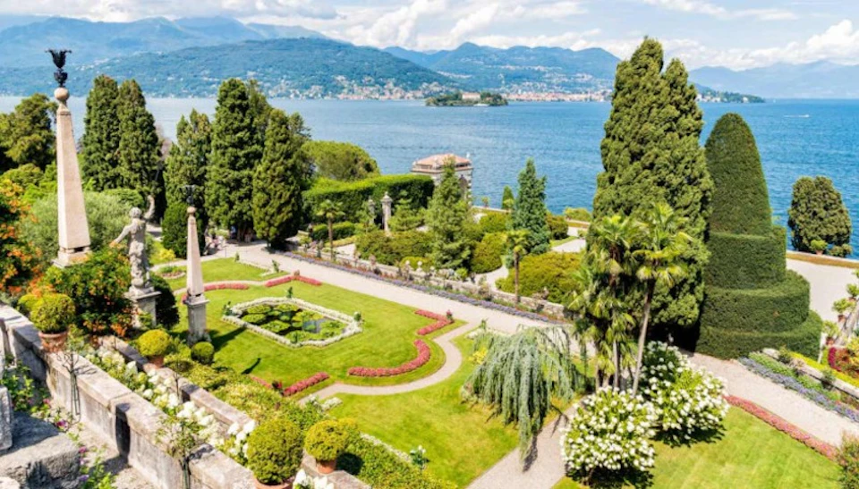 Manicured garden with shaped hedges, paths, and flowers overlooking a large lake with mountains in the background.