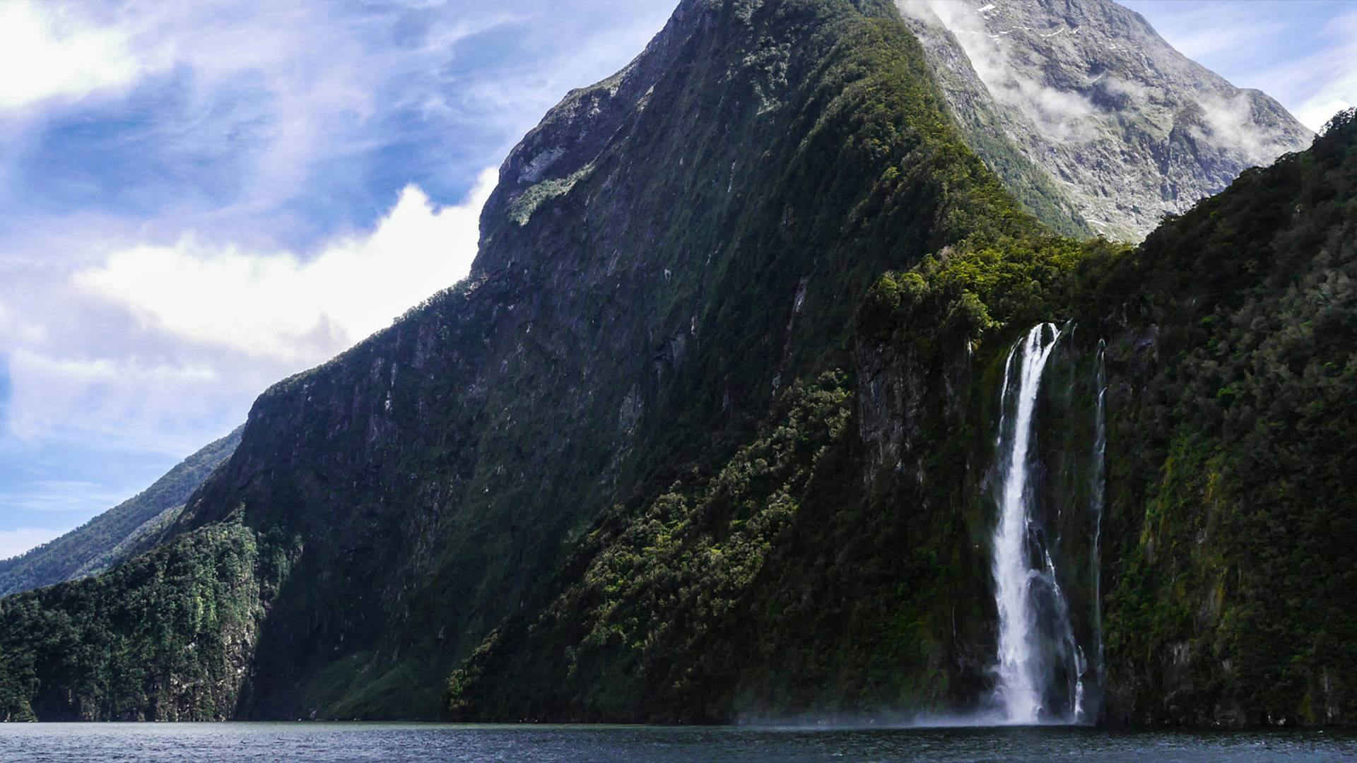 Uma cascata alta que desce em cascata por uma montanha íngreme e arborizada até uma massa de água tranquila, com um céu azul parcialmente nublado.