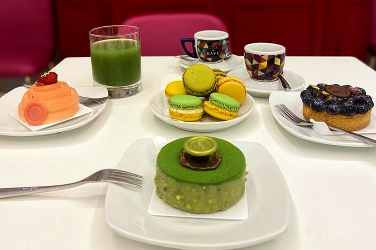 配有绿色蛋糕、各种马卡龙、五颜六色的咖啡杯、绿色果汁和叉子的桌子。 后面是红色的椅子。