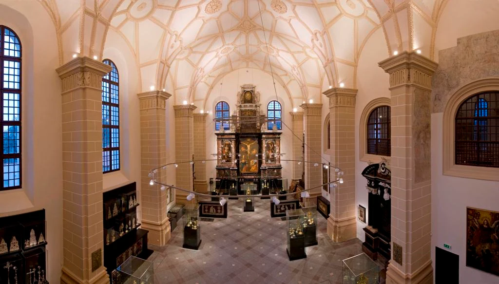 A grand hall with high, ornate ceilings, an altar at the back, display cases, and artwork adorning the walls.