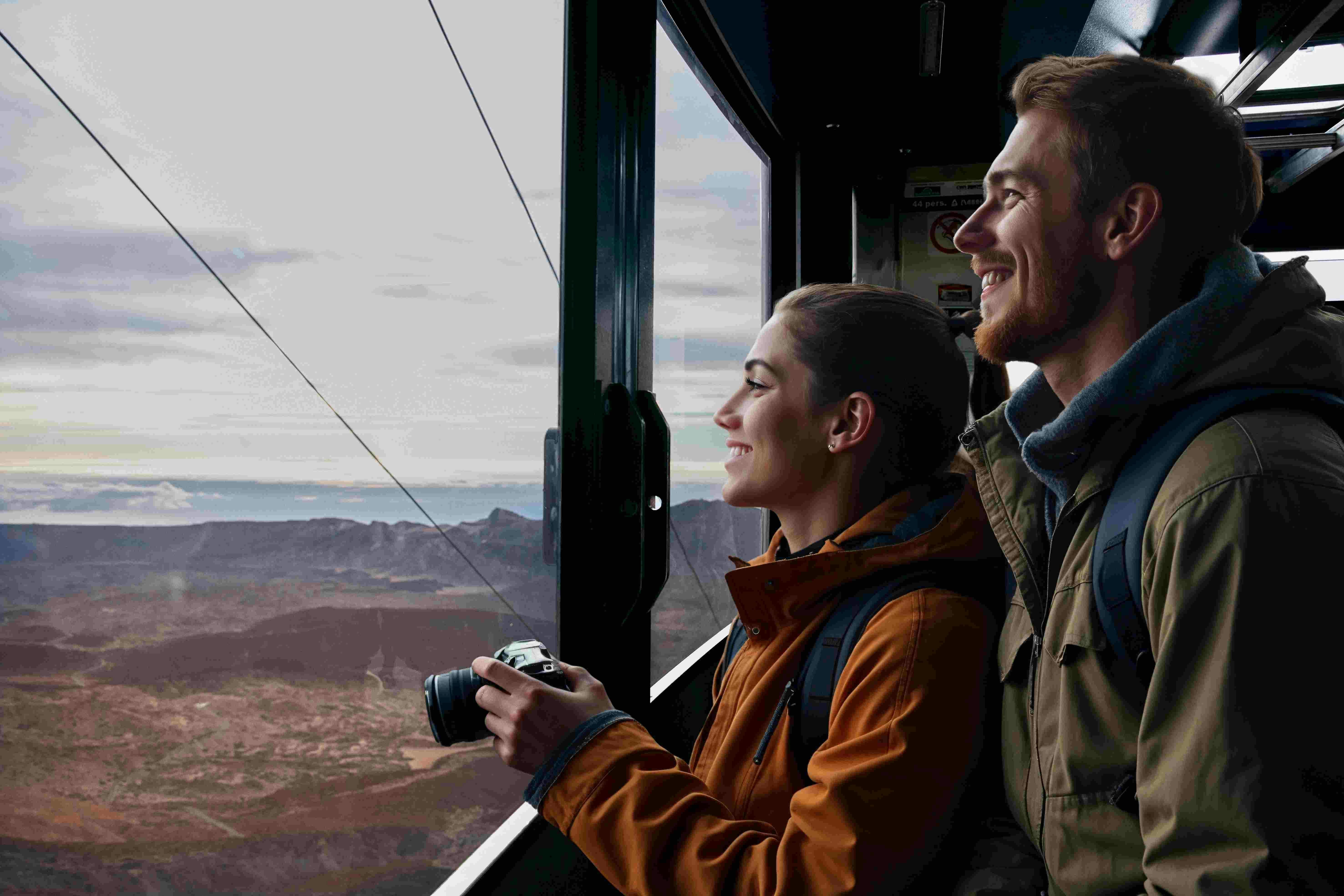 A smiling man and woman ride a cable car, looking out the window at mountain scenery. The woman holds a camera, both wear jackets.