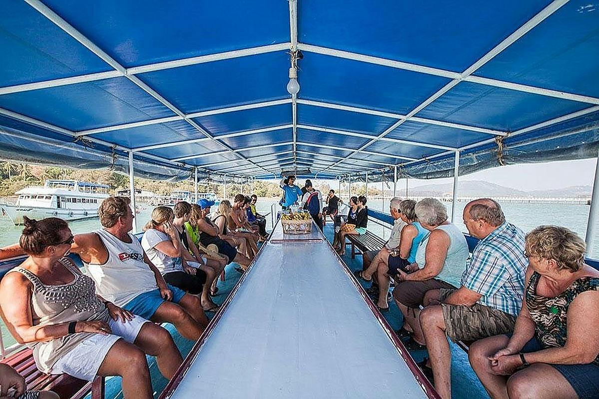 People sitting on benches under a blue canopy on a boat, with a white table in the center and a scenic view in the background.