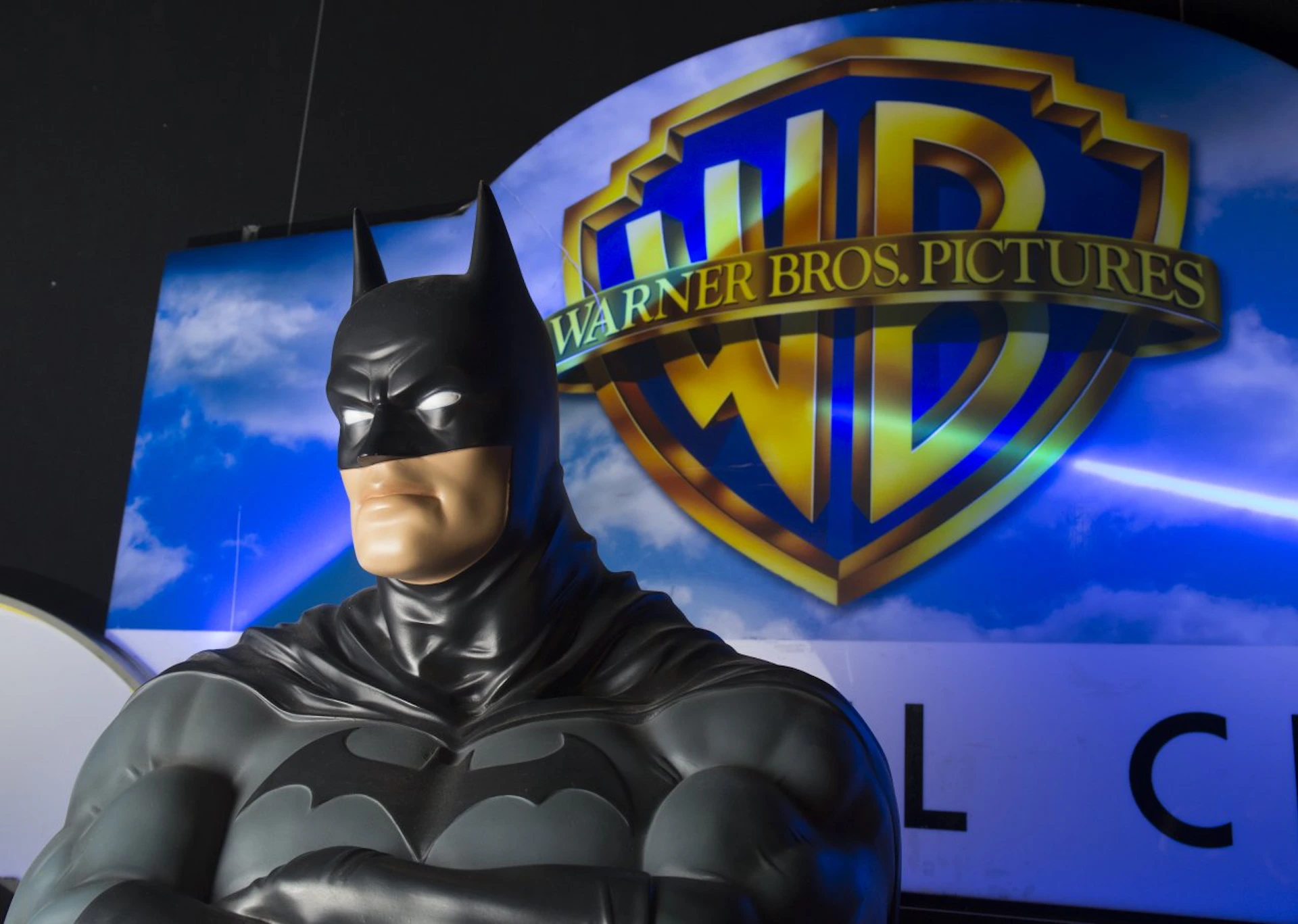 Statue of Batman with crossed arms in front of a Warner Bros. logo and a sky background.