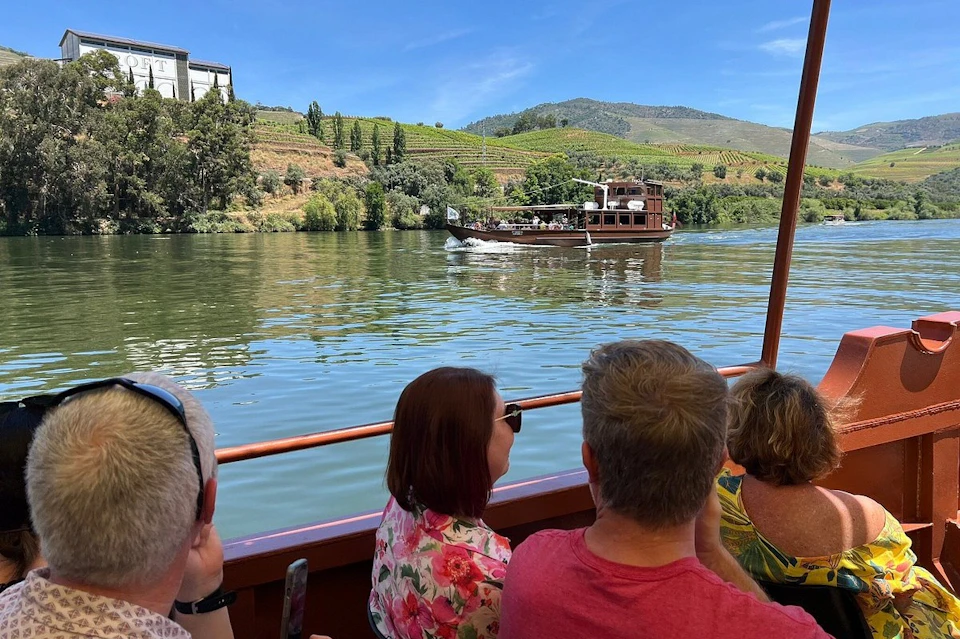 People on a boat look out at another boat on a river, with vineyards and hills in the background under a clear blue sky.