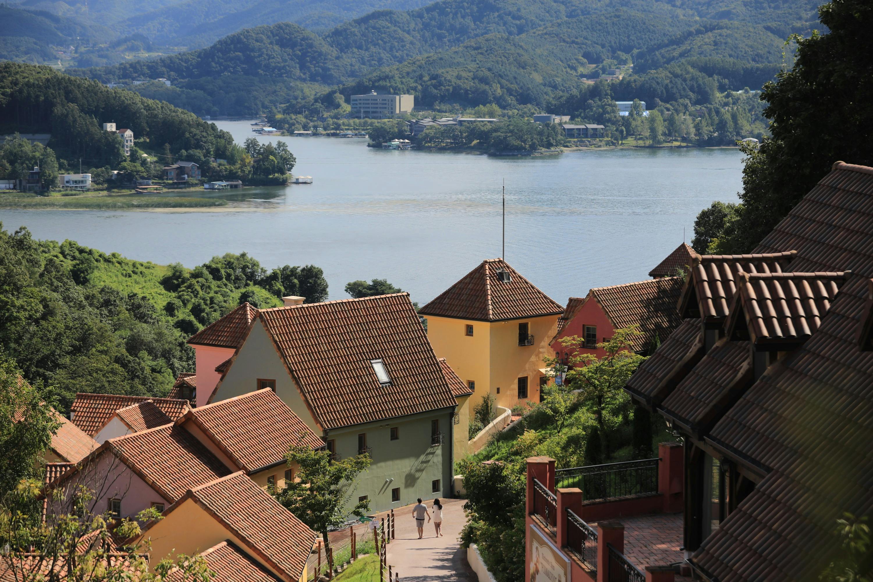 A scenic view of colorful houses with red roofs on a hill, overlooking a lake surrounded by green hills and trees.