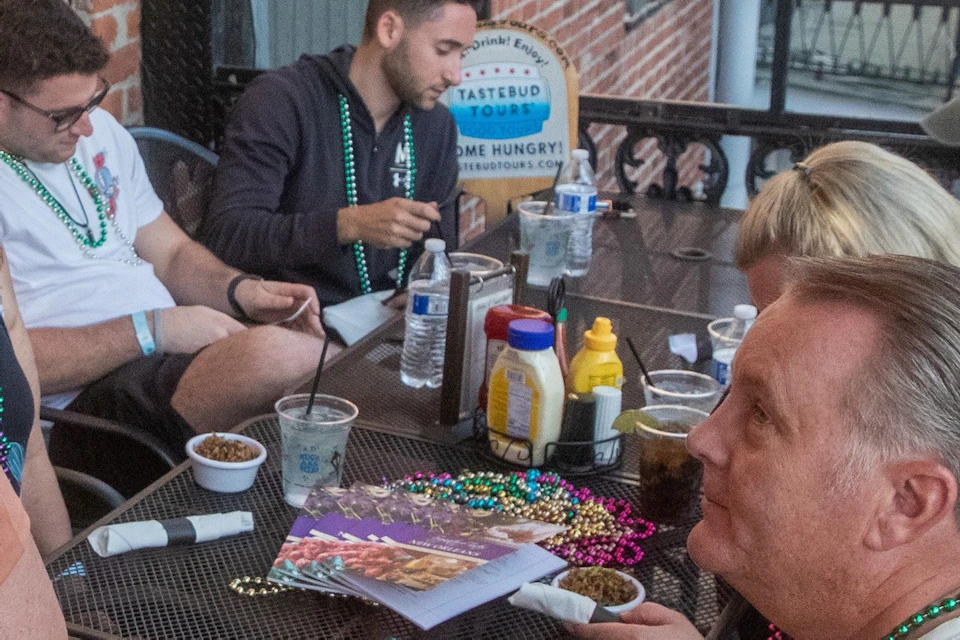 A group of people sitting at a table outdoors, wearing beads, with food and drinks on the table, near a brick building.