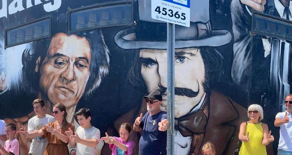 A group of people in front of a mural depicting two men with intense expressions. They are posing playfully and pointing at the camera.