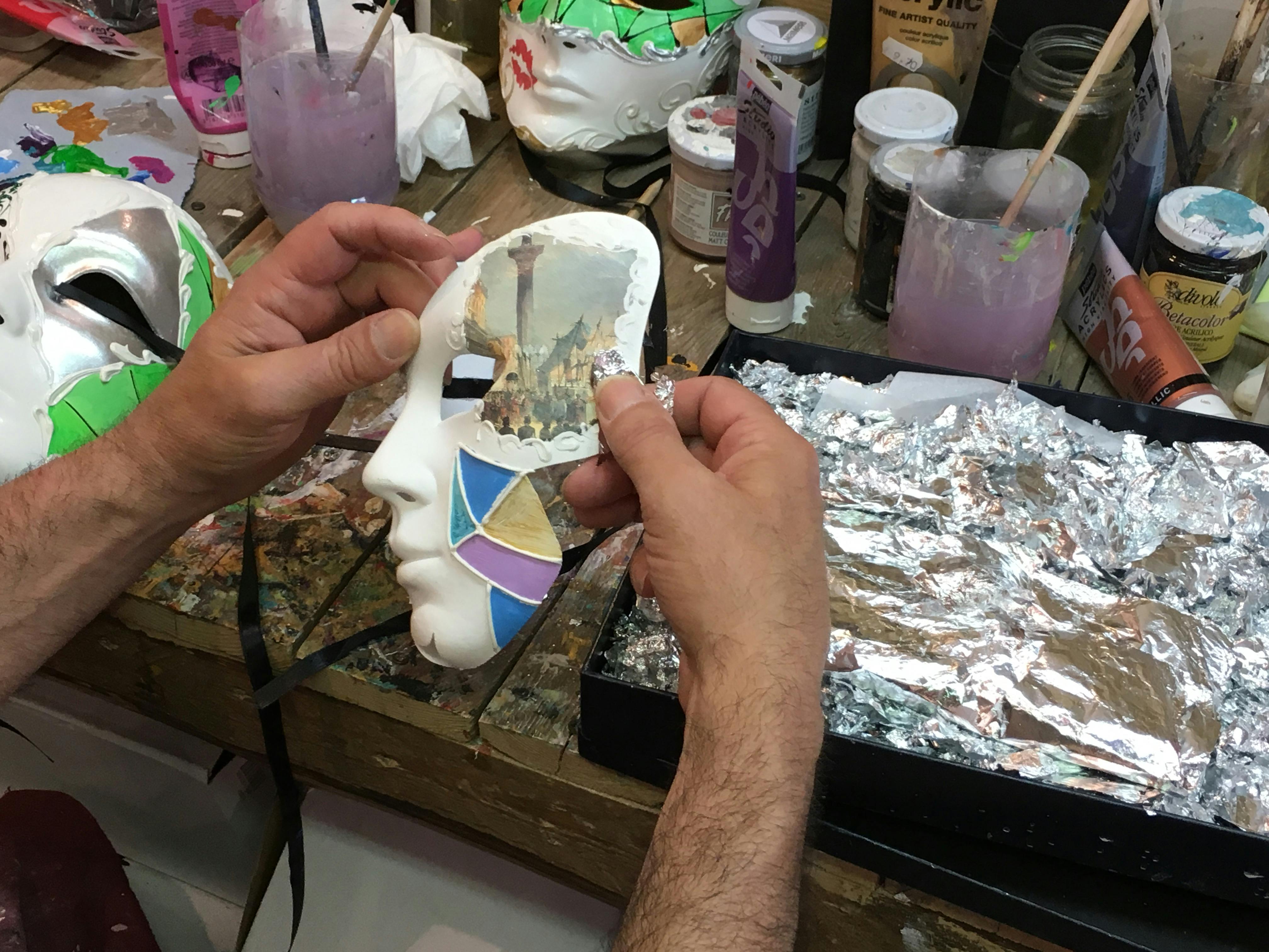 페인트 용품, 브러시, 호일이 있는 어수선한 작업 공간 위에 장식된 마스크를 들고 있는 손.