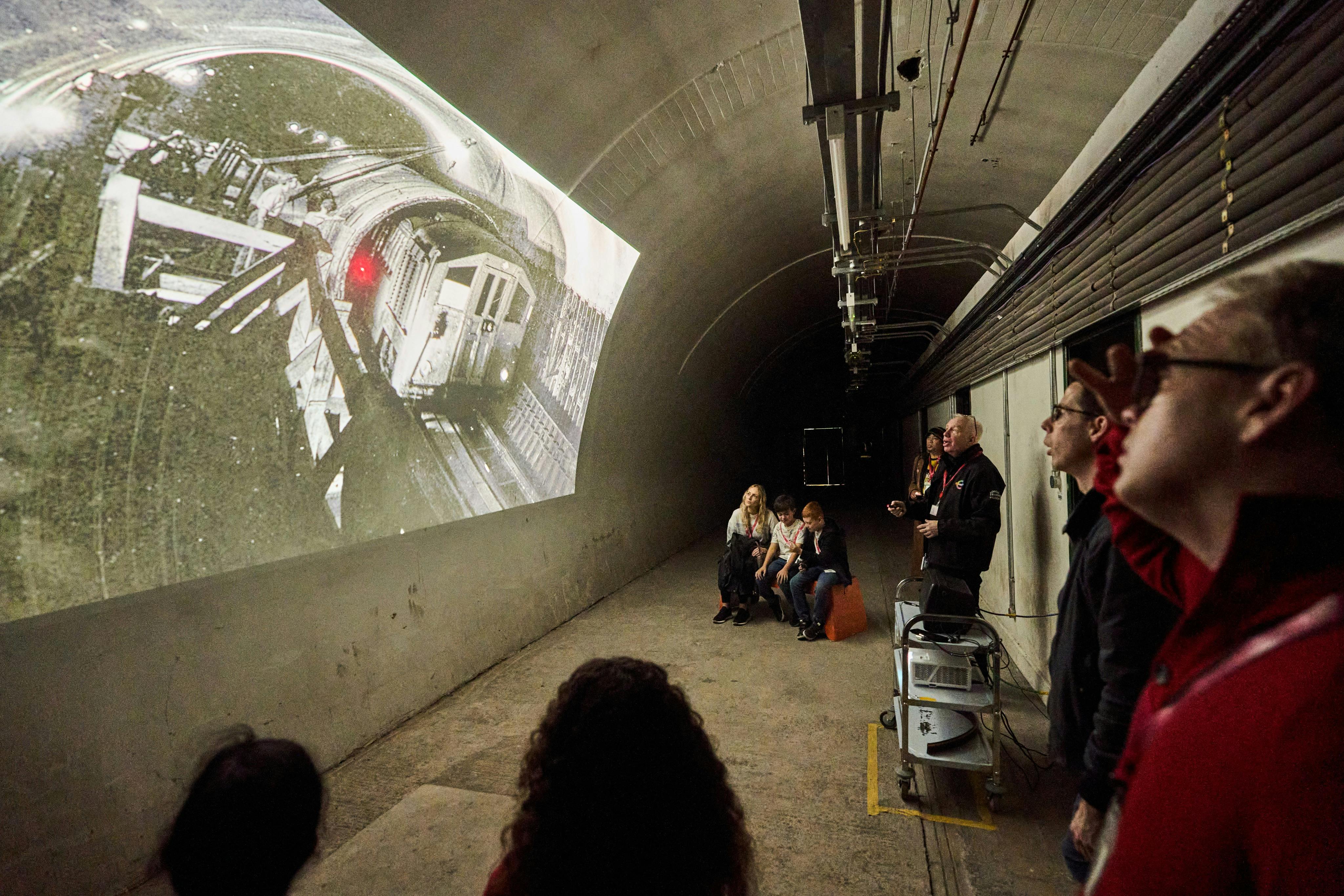 Mensen verzamelden zich in een tunnel en keken naar een geprojecteerd beeld van een oude trein op de muur, met een spreker die een presentatie gaf.