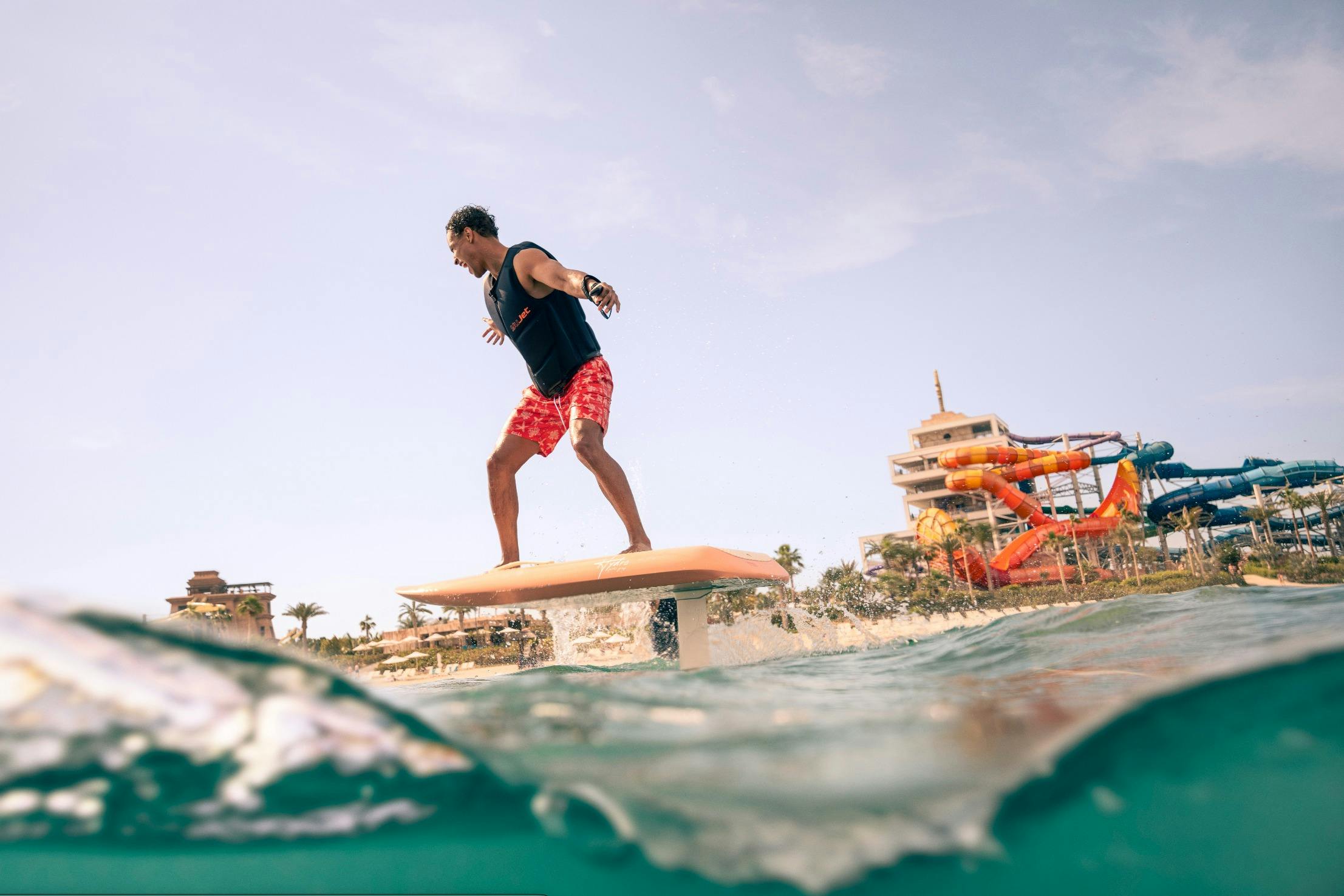 Человек балансирует на доске для серфинга на подводных крыльях над водой рядом с пляжем, на заднем плане видны здания и пальмы.