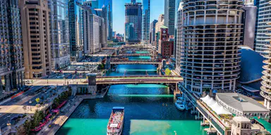 Una veduta aerea di un paesaggio urbano con edifici alti, ponti multipli e un fiume con una barca che trasporta persone.