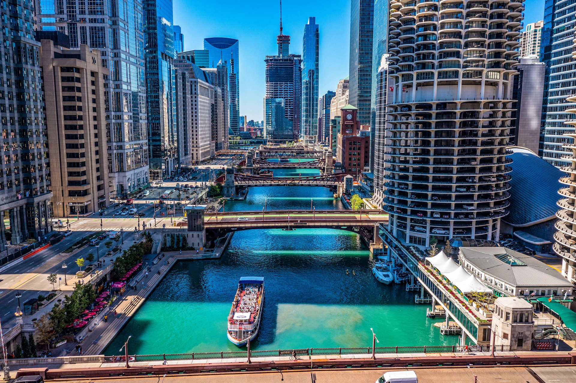 An aerial view of a cityscape with tall buildings, multiple bridges, and a river with a boat carrying people.