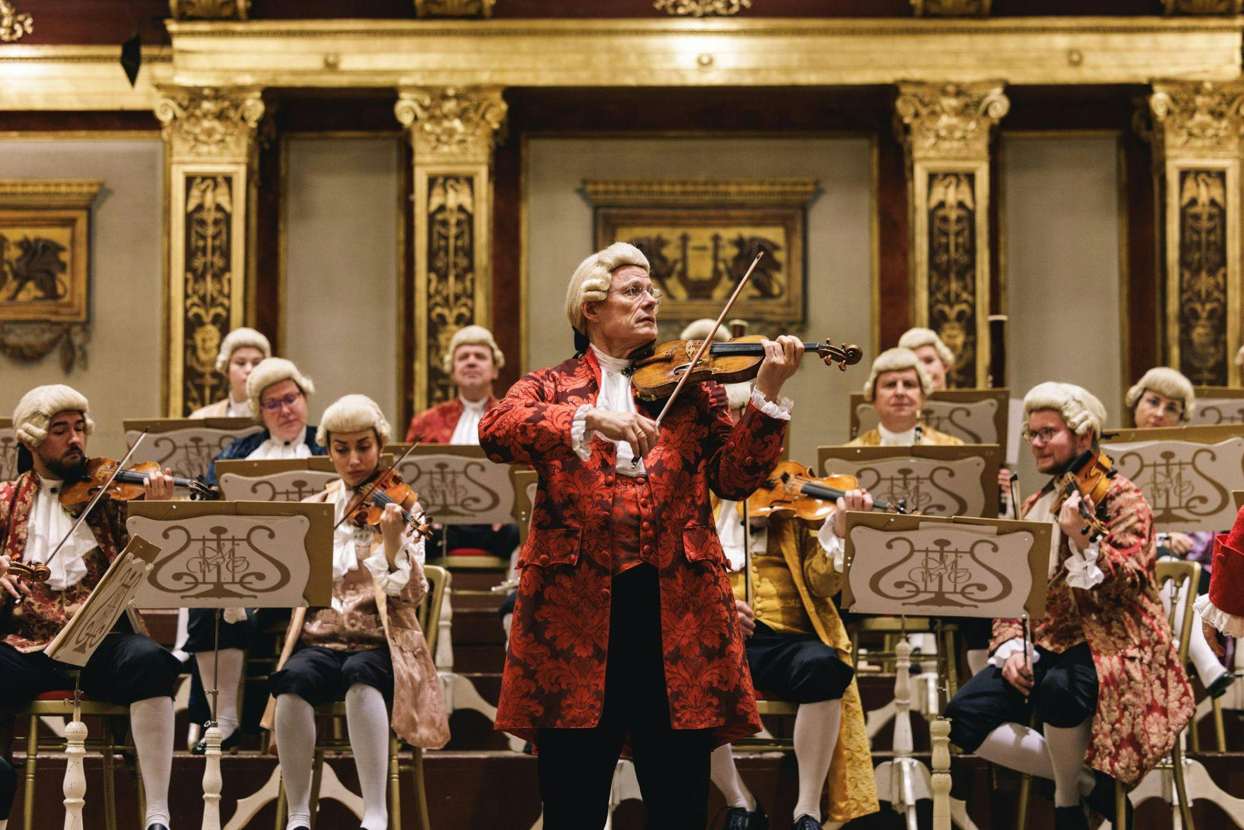 Musicians in 18th-century attire perform in a gilded concert hall, with a violinist in the foreground.