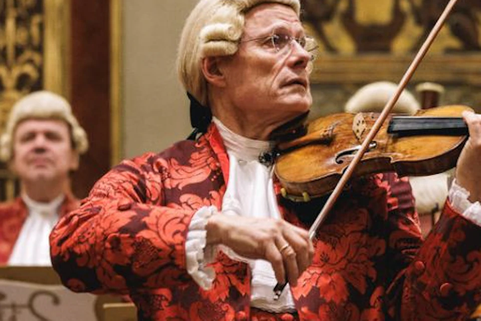 Musicians in 18th-century attire perform in a gilded concert hall, with a violinist in the foreground.