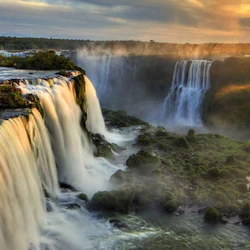 Majestic waterfalls cascade down rocky cliffs into a misty, lush green landscape under a dramatic sky at sunset.