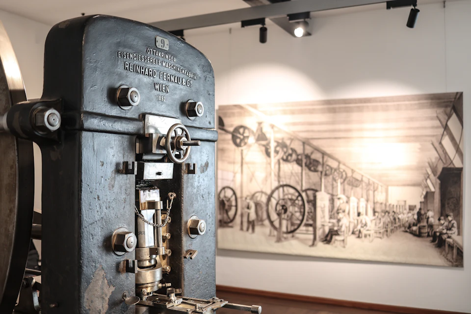 A vintage industrial machine displayed in a museum, with a historical illustration of machinery in the background.