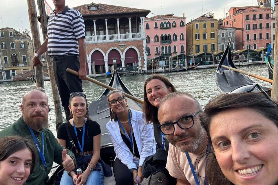 A group of seven people on a gondola, with a gondolier standing at the back. Venetian buildings line the canal in the background.