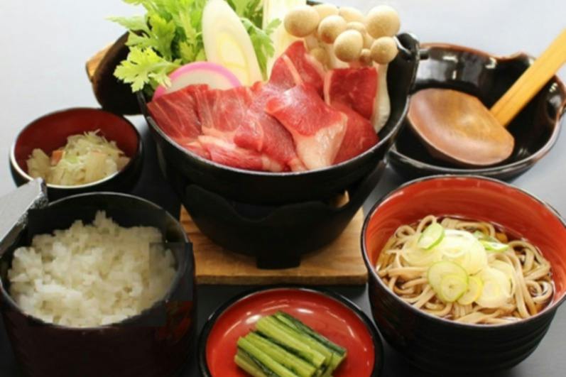 A Japanese hot pot meal with raw beef, mushrooms, and vegetables, served alongside rice, noodles, and pickled greens.
