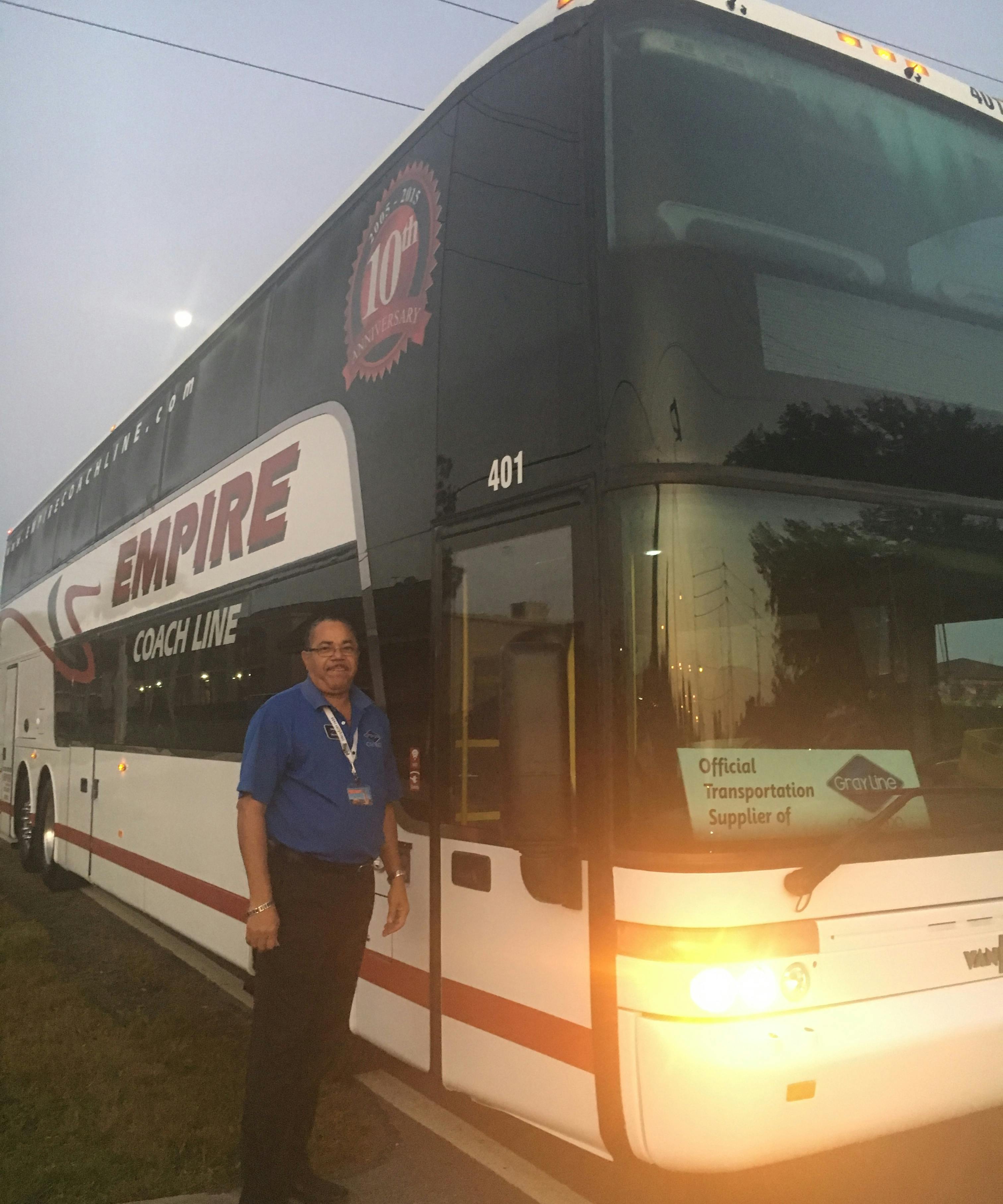 A person wearing a blue shirt stands beside a double-decker Empire Coach Line bus. A sign reads, "Official Transportation Supplier."