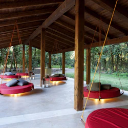 Täckt utomhuslounge med upphängda runda röda dagbäddar, grå kuddar och lyktor. Omgiven av gräsyta och träd.