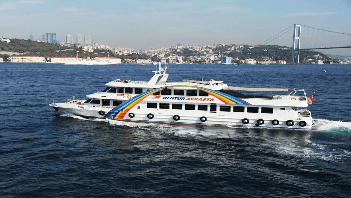 「Dentur Avrasya」というレインボーデザインのフェリーが、街並みと橋を背景に水上を航行しています。