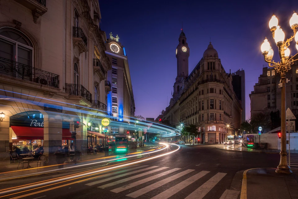 Nighttime city street scene with light trails from vehicles, historic buildings, and a clock tower under a twilight sky.