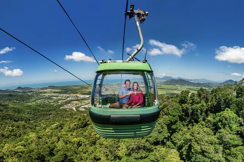 A couple sits inside a green cable car, suspended over a lush forest with a scenic landscape in the background.