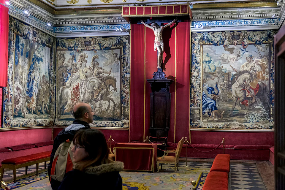 Two people observing an ornate room with tapestries, a crucifix, and red velvet curtains.
