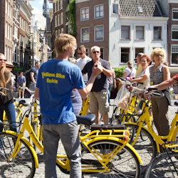 En grupp människor med gula cyklar lyssnar på en guide som bär en blå "Yellow Bike Guide" -skjorta på ett torg med höga byggnader.