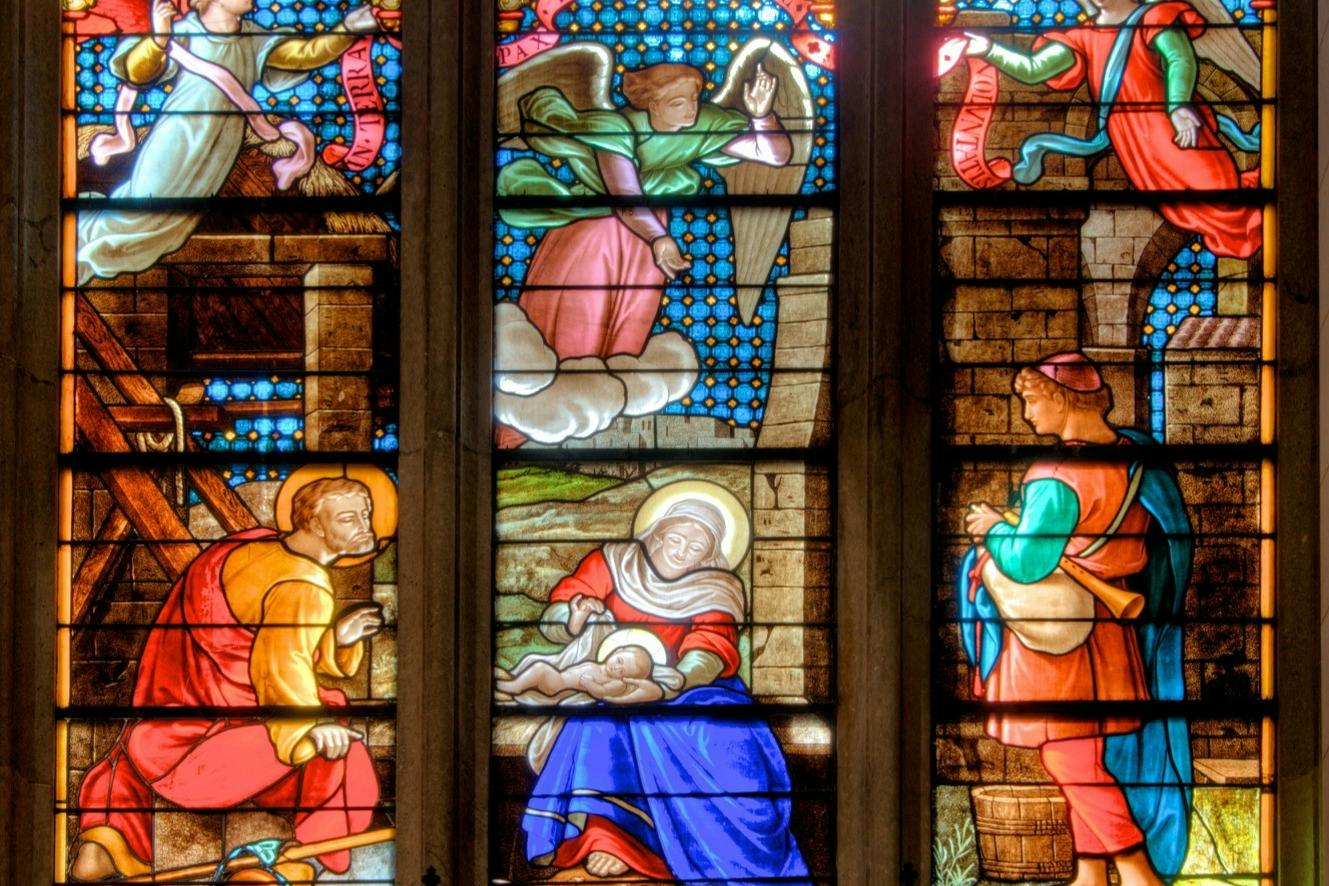 A stained glass window depicting a nativity scene with Mary, Joseph, baby Jesus, an angel, and shepherds in vibrant colors.