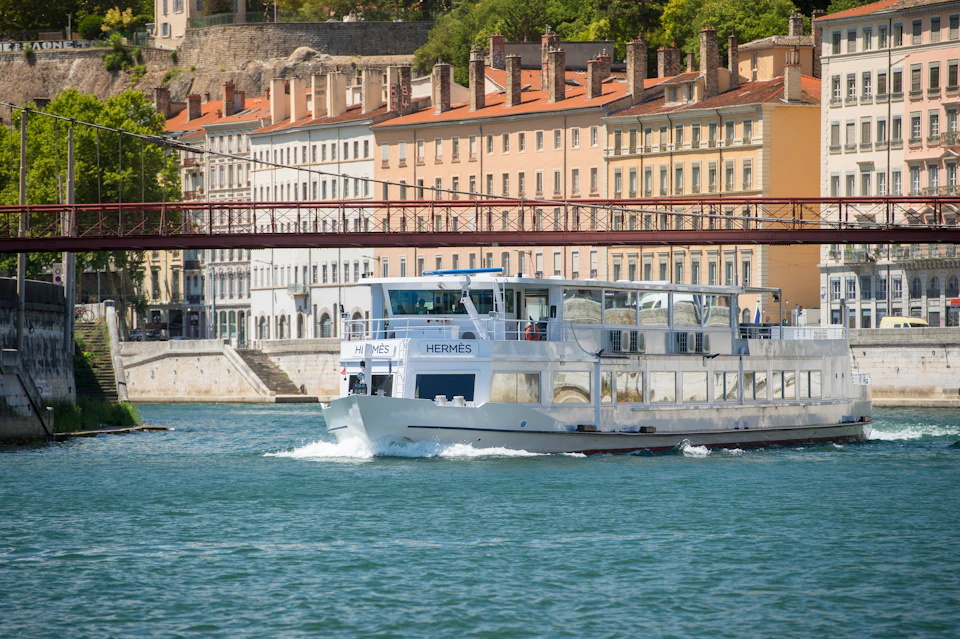 A white river cruise boat labeled "Hermès" sails on a river with historic buildings and a red bridge in the background.