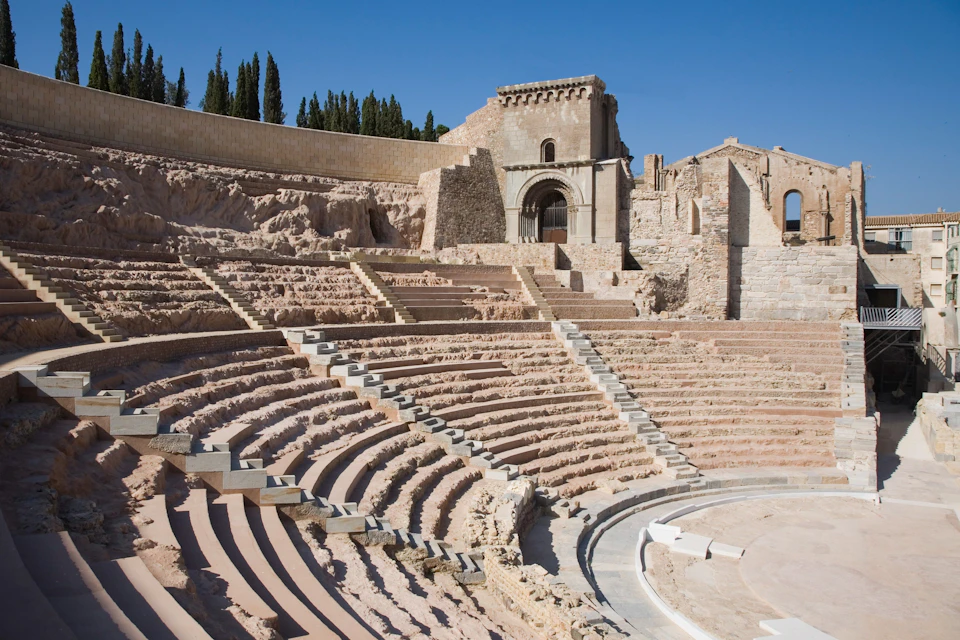 Ancient Roman amphitheater with tiered seating and weathered stone architecture under a clear blue sky.