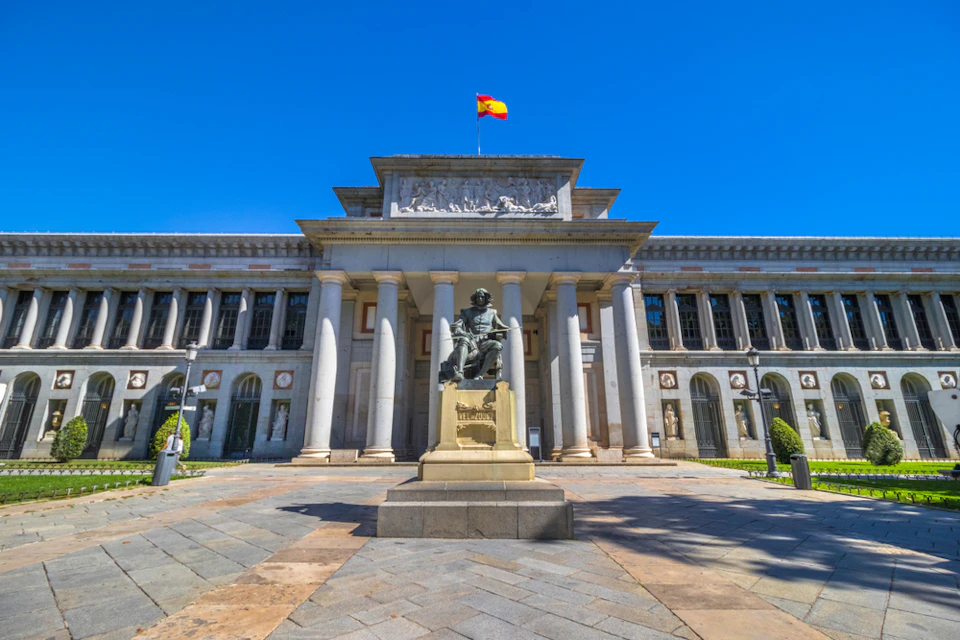 Grand neoclassical building with columns and a statue in front. A Spanish flag flies on top against a clear blue sky.