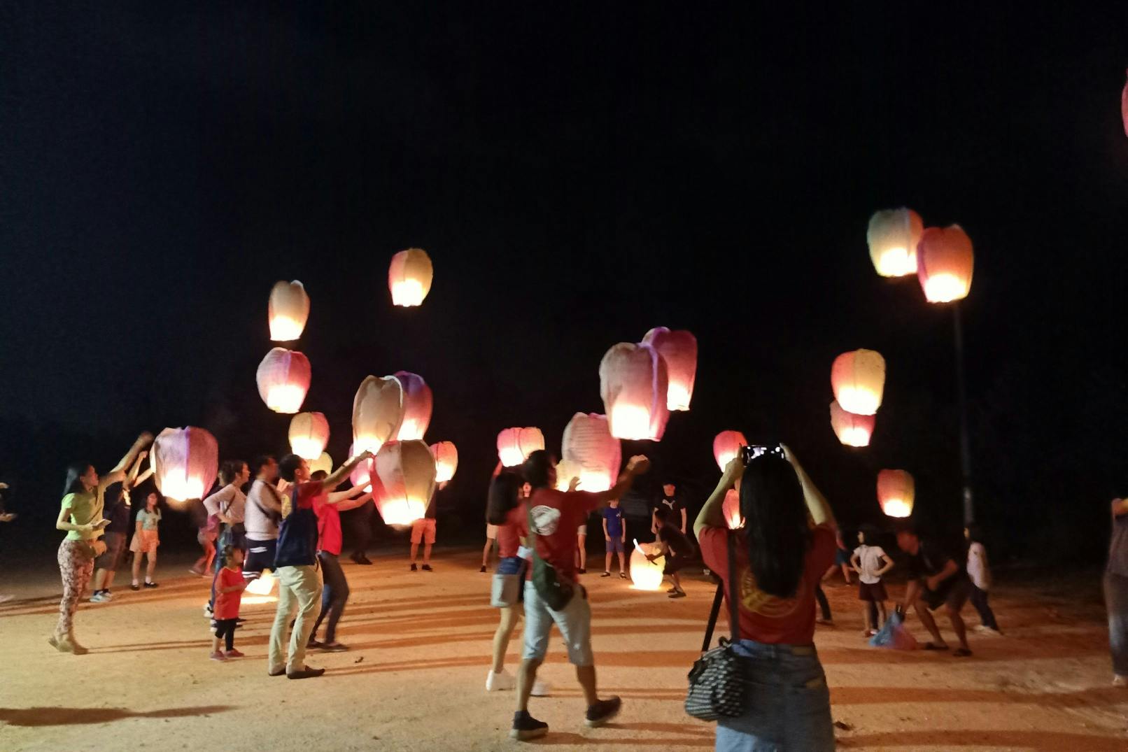 People releasing illuminated paper lanterns into the night sky, with a crowd watching and participating.