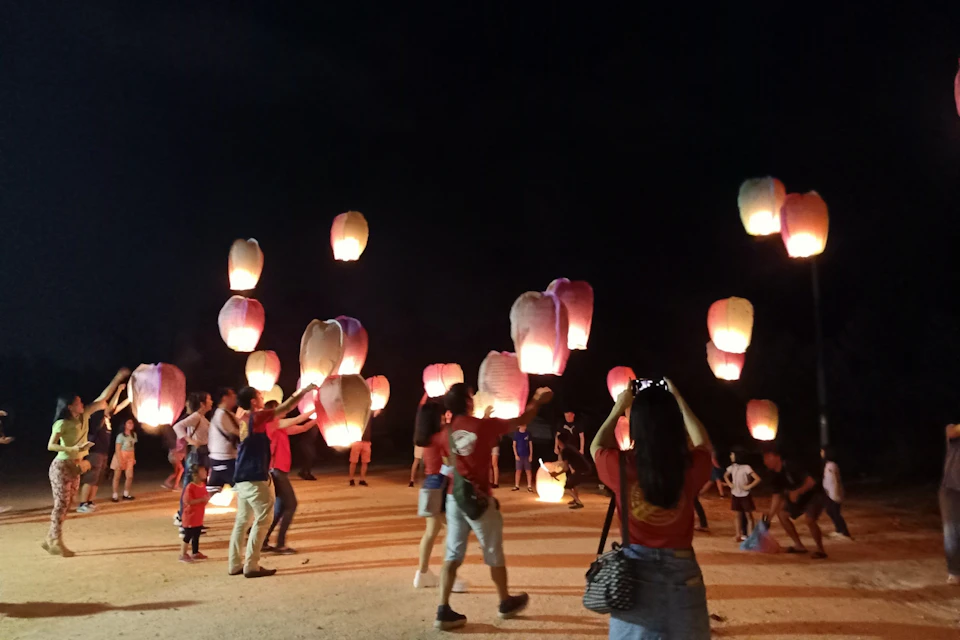 People releasing illuminated paper lanterns into the night sky, with a crowd watching and participating.