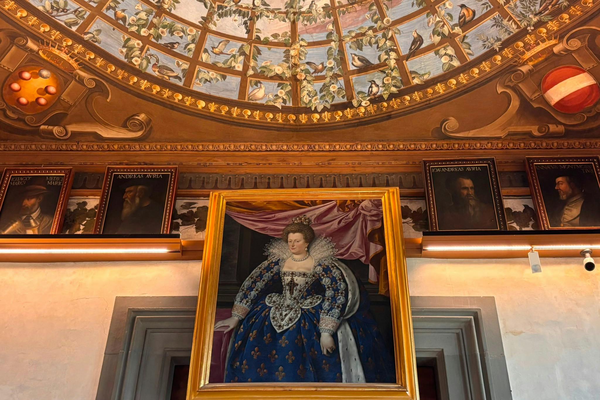 Um retrato emoldurado de uma mulher com trajes majestosos, sob um teto abobadado ornamentado com pássaros e folhagem pintados.