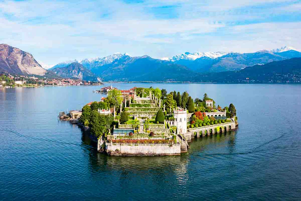 Lush, terraced gardens and buildings on a small island surrounded by calm water, with mountains in the background under a clear sky.