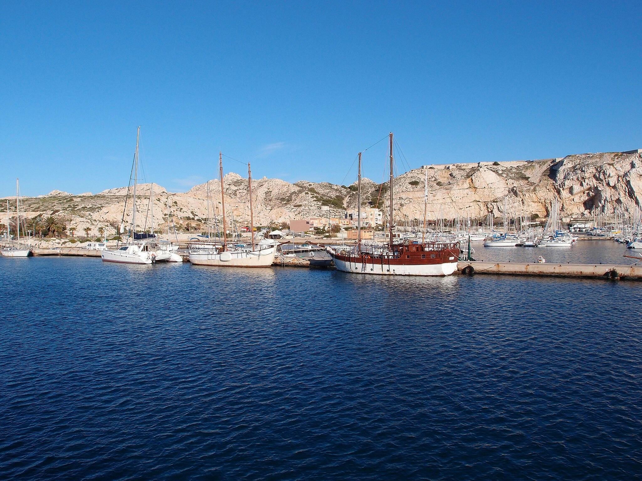Kilka łodzi i żaglowców zakotwiczonych w doku z kamienistymi wzgórzami i budynkami w tle pod czystym błękitnym niebem.
