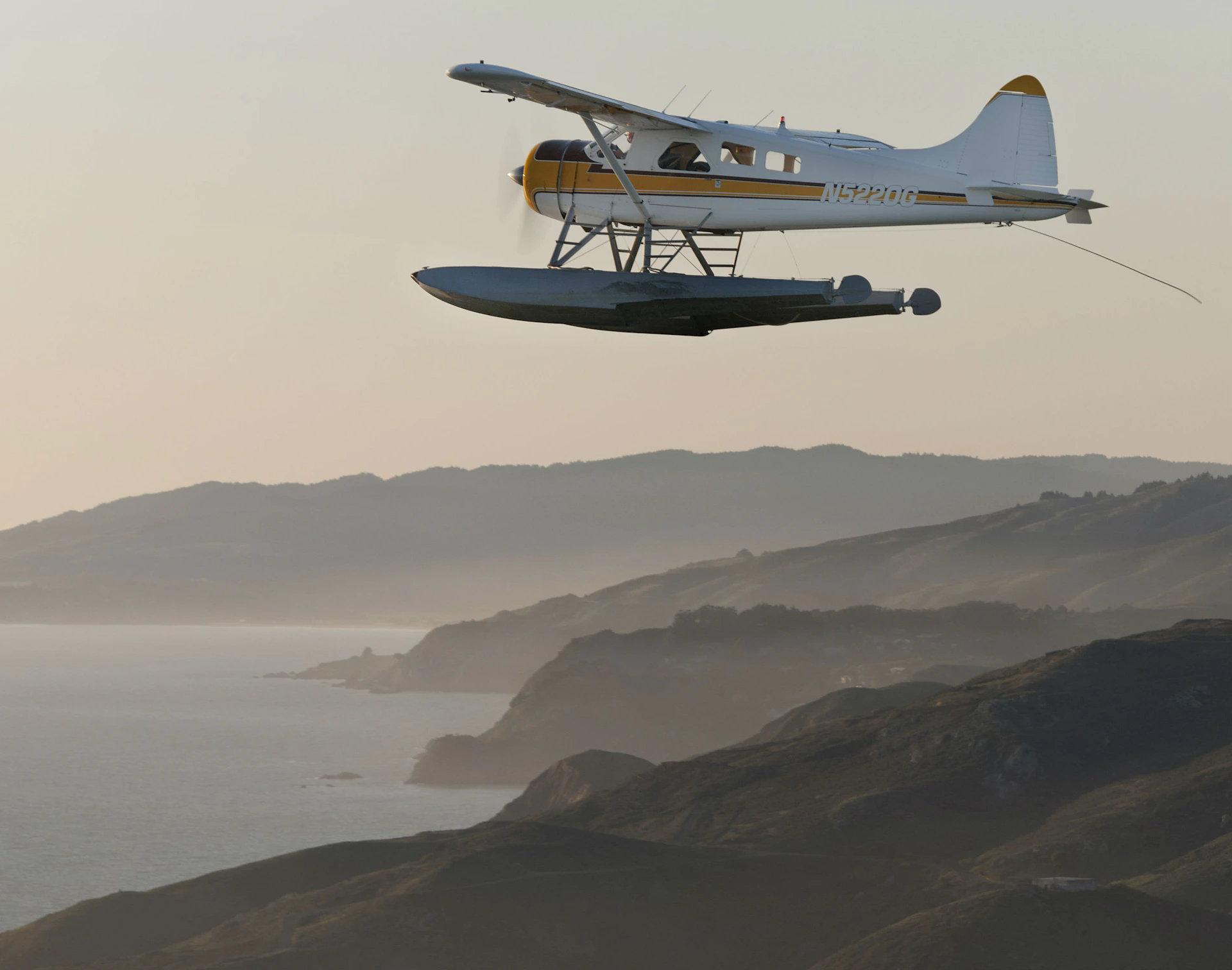 A small seaplane with floats flies over a coastal landscape at sunset, with misty hills and the ocean in the background.