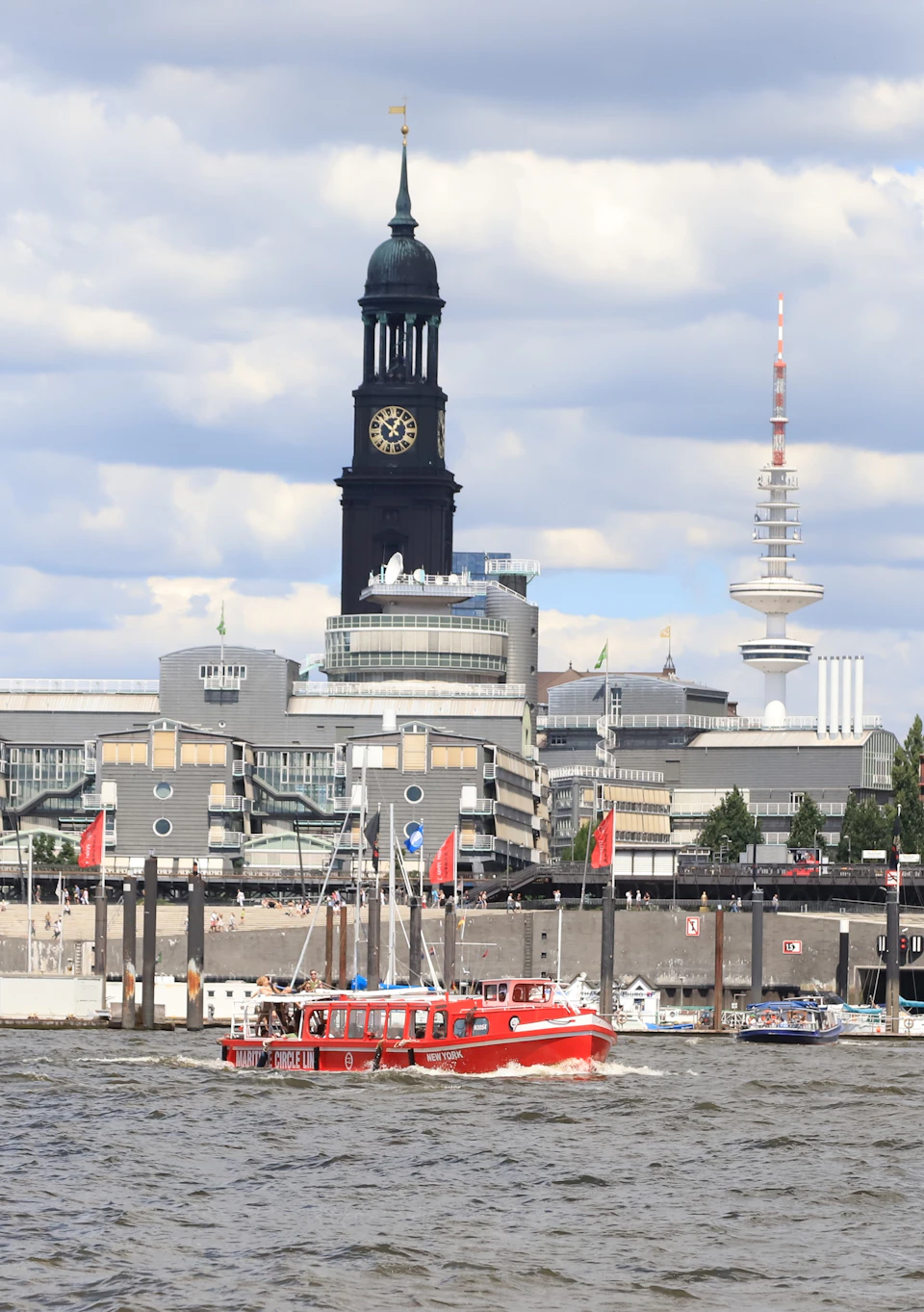 A red tour boat sails on a river with a cityscape in the background, featuring a large clock tower and modern buildings under a cloudy sky.