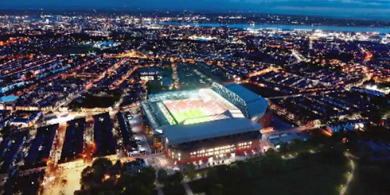 Widok z lotu ptaka na jasno oświetlony stadion pośród miejskich świateł o zmierzchu, z otaczającymi go obszarami mieszkalnymi i miejskimi.