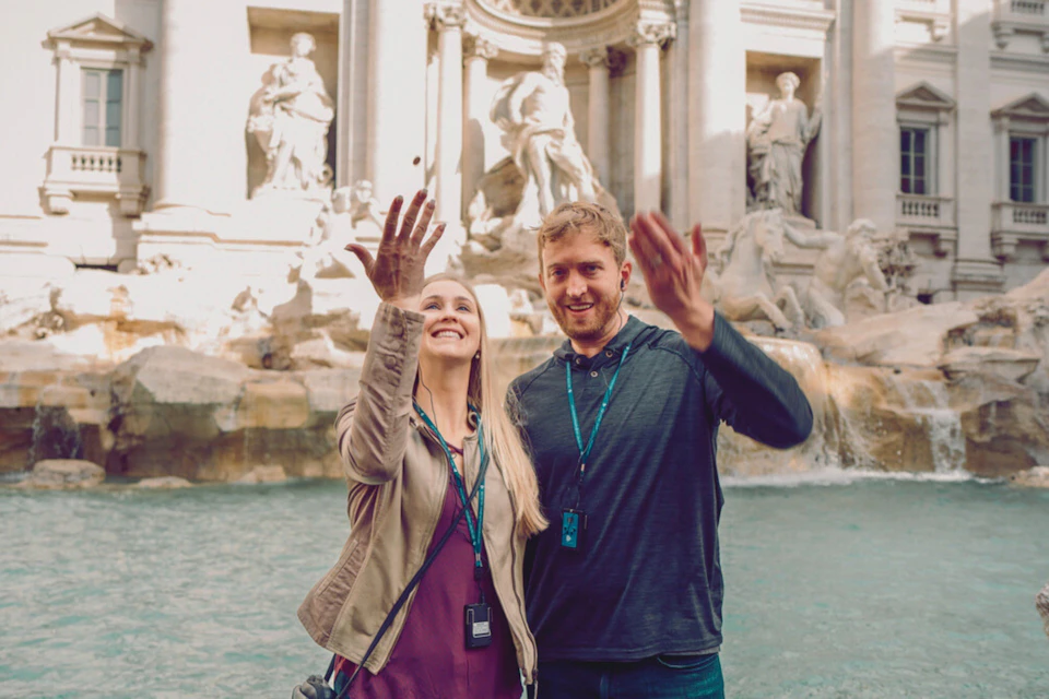 A smiling couple tosses coins into the Trevi Fountain in front of ornate sculptures on a sunny day.