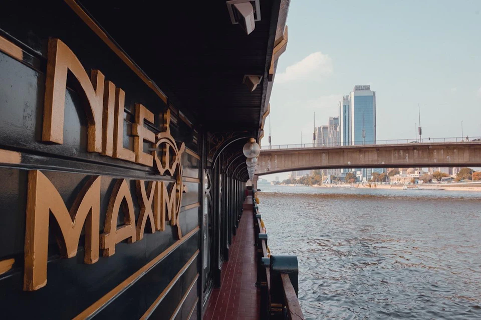 A docked boat with 'Nile Maxima' on its side, a river, a bridge, and city buildings in the background under a clear sky.