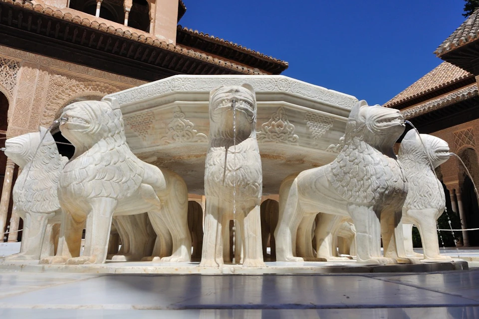 A stone fountain supported by intricately carved lion statues, under a clear blue sky with part of a building visible.