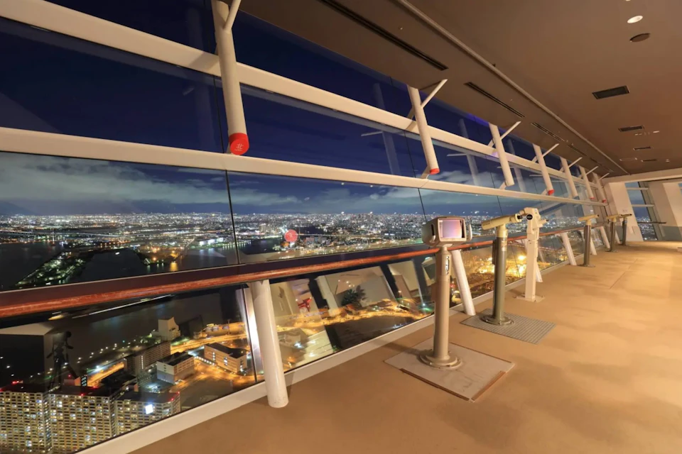 Observation deck with telescopes overlooking a brightly lit cityscape at night through large glass windows.
