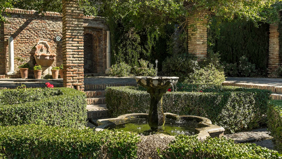 A stone fountain surrounded by neatly trimmed hedges and brick arches in a lush garden.