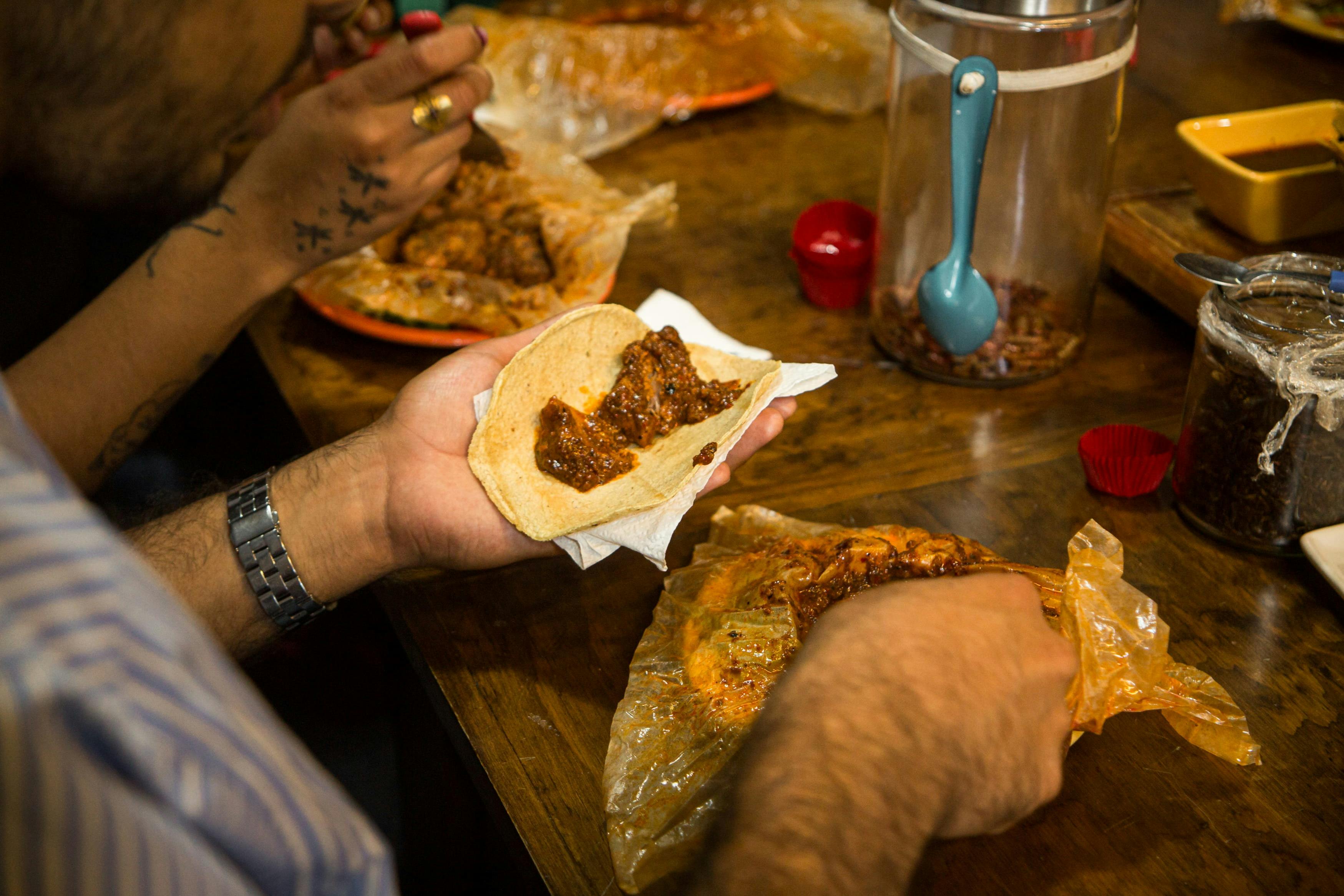 Zwei Personen bereiten Tacos mit Fleisch und Soße an einem Holztisch zu, mit Lebensmittelbehältern und Tellern in der Szene.